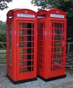 two red telephone booths sitting next to each other on top of a gravel field with trees in the background