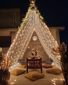 a tent with lights and pillows on the floor in front of a table surrounded by potted plants