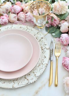 the table is set with pink flowers and gold place settings for two people to sit at