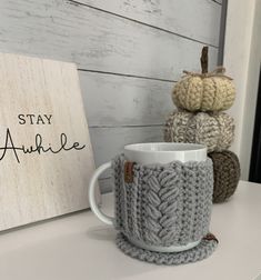 two knitted mug cozys sitting next to a sign that says stay awhile on it