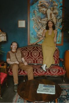 a man and woman sitting on a couch in front of a wall with paintings behind them