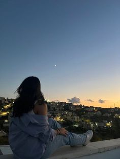 a woman sitting on top of a roof looking at the city lights in the distance