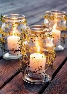 some candles are sitting in glass jars on a wooden table with flowers and greenery