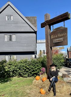 a woman sitting on top of hay next to a sign and pumpkins in front of a house