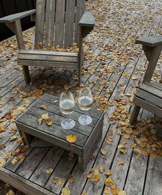 two wooden chairs sitting on top of a wooden deck covered in leaves and water glasses