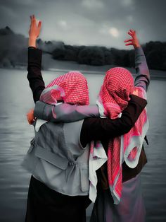 two women wearing headscarves and scarves are standing in front of a body of water