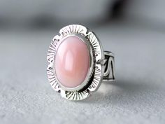 Bold but dainty Pink Conch Silver ring on a unique silver band that has been handmade using scrap 925 Sterling Silver. The band has been fused, oxidized then lightly polished. At its widest the band is 3/8" then tapers to 1/8". The oval cabochon is 14mm x 10mm and is in a bezel setting accented with stamped, cut out square wire. Add a touch of Coastal Cowgirl attitude to your outfit with our simple, but elegant Pink Conch Silver Statement Ring. Whether you're shopping for yourself or are searchi Pink Oval Jewelry Stamped 925, Oval Cabochon Opal Ring With Bezel Setting As Gift, Oval Pink Opal Ring For Gift, Pink Opal Ring In Sterling Silver For Anniversary, Pink Oval Jewelry With Bezel Setting, Oval Pink Opal Ring Gift, Pink Oval Opal Ring For Gift, Pink Sterling Silver Opal Ring For Anniversary, Pink Opal Rings As A Gift