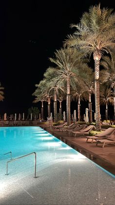 an empty swimming pool with lounge chairs and palm trees in the background at night time