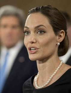 a woman in a black dress with pearls on her neck and two men in suits behind her