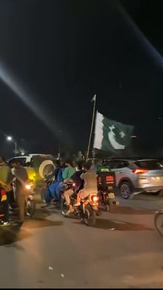 motorcyclists are riding down the street at night with flags flying in the background