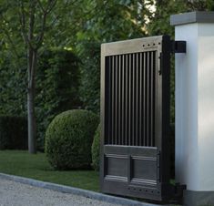 an open gate in front of a white wall and some bushes on the other side