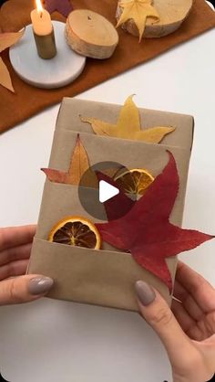 a person holding a piece of paper with oranges and leaves on it in front of a candle