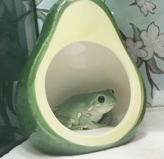 a green frog sitting inside of an avocado shaped vase with flowers on the wall behind it