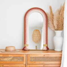 a wooden dresser topped with a mirror next to a white vase filled with dry grass