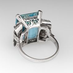 This striking vintage cocktail ring is centered with an emerald cut aquamarine, weighing 10.94 carats, in a four-prong setting. The shoulders are each accented with a bow design with milgrain details and is bead set with nine (9) round single cut diamonds. The ring measures 16.8mm at the top, rises 8.9mm above the finger, tapering to 1.9mm wide and 0.9mm thick at the base of the shank. This ring is currently a size 5. There is a tiny nick on the face of the stone. Vintage Cocktail Ring, Diamond Cocktail Ring, Aquamarine Jewelry, Diamond Cocktail Rings, Bead Set, Vintage Cocktail, Bow Design, March Birth Stone, Cocktail Ring