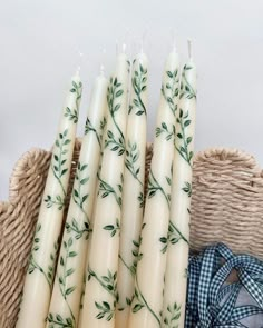 four candles with green leaves on them sitting in a basket next to a blue and white checkered napkin