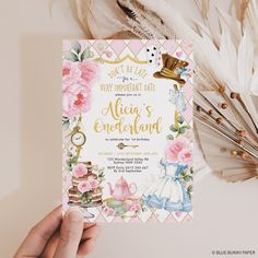 a person holding up a pink and gold princess birthday party card next to some feathers