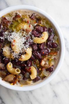 a white bowl filled with pasta and beans on top of a marble counter next to a spoon