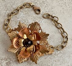 "The centerpiece for this fun layered flower bracelet is a construction of 3 different shaped flower petal stampings riveted to a very pretty vintage brass filigree (see photo of back).  A large root beer rhinestone is featured at the center of the flower. The filigree is about 2\" wide by around 3\" long and curved to fit the wrist; textured antique gold chain makes up the rest of the length. It fastens with a medium sized lobster clasp that can hook anywhere on the other side, so may be worn f Vintage Metal Jewelry With Flower Charm, Vintage Gold Flower Shaped Jewelry, Adjustable Vintage Flower Shaped Jewelry, Vintage Copper Bracelet Jewelry, Vintage Gold Bracelets With Copper, Vintage Gold Bracelets With Copper Material, Vintage Brass Jewelry With Flower Charm, Vintage Flower Bracelets For Jewelry Making, Vintage Copper Bracelet