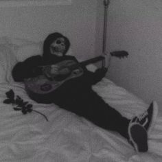 a person in a black hoodie playing an acoustic guitar on a bed with white sheets