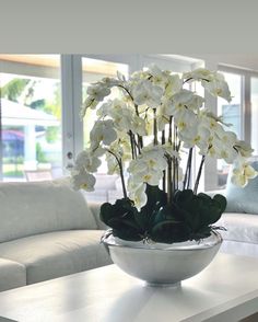 a white flower arrangement in a bowl on a table with couches and windows behind it