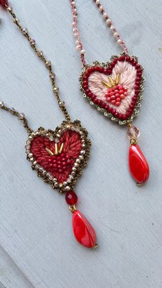 two red and white heart shaped pendants hanging from chains on a wooden table with beads