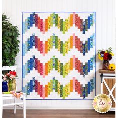 a colorful quilt hanging on the wall next to a white chair and potted plant