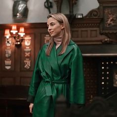 a woman in a green trench coat standing next to a fireplace