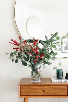 a vase filled with red berries and greenery on top of a wooden table next to a mirror