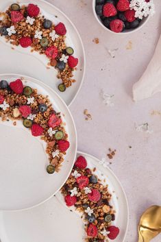 three white plates topped with granola and berries next to a gold spoon on a table