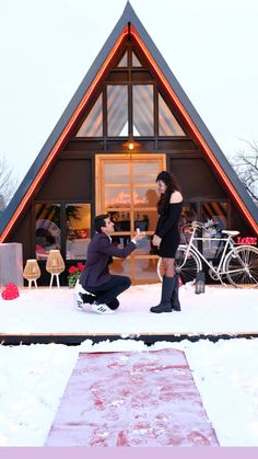 a man kneeling down next to a woman in front of a house with snow on the ground