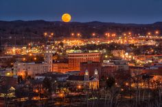 the full moon is seen over a city at night with buildings lit up and trees in the foreground