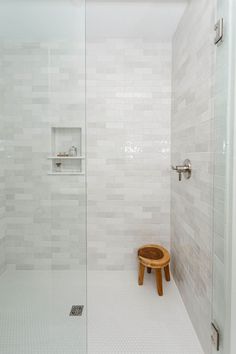 a walk in shower sitting next to a white tiled wall and floor with a wooden stool