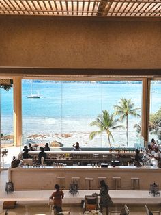 people are sitting at tables in front of large windows overlooking the water and beach with palm trees