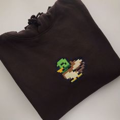 The Stardew Duck Hoodie Apparel by petimint is a stylish folded black sweatshirt, featuring a pixelated image of a duck on the chest with a green head, yellow beak, and brown and white body. Perfect for gaming enthusiasts, this piece of apparel is neatly arranged on a light grey surface. Green Casual Hoodie With Custom Embroidery, Casual Green Hoodie With Custom Embroidery, Duck Hoodie, Embroidered Design, Military Green, Ethical Fashion, Slow Fashion, Original Design, Original Designs