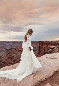 a woman standing on top of a cliff wearing a long white dress and looking at the sky