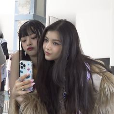 two girls are looking at their cell phones while standing in front of a mirror and one girl is wearing a fur coat