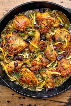 a skillet filled with chicken, onions and olives on top of a wooden table