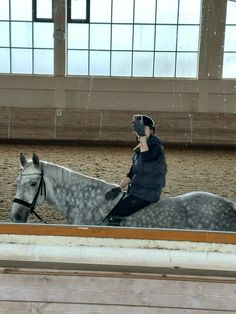 a woman riding on the back of a gray horse in an indoor arena with large windows