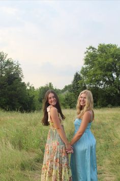 two women standing next to each other in a field with tall grass and trees behind them