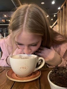 a girl drinking from a coffee cup on top of a saucer next to a brownie