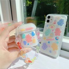 a hand holding an iphone case with charms attached to it, next to a window