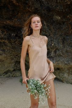 a woman standing on the beach with flowers in her hand and wearing a tan dress