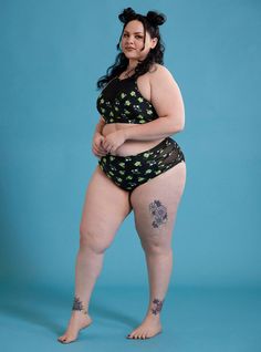 a woman in a black and green swimsuit with tattoos on her legs, standing against a blue background