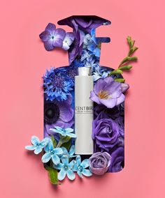 a bottle of lotion sitting on top of purple and blue flowers next to a pink background