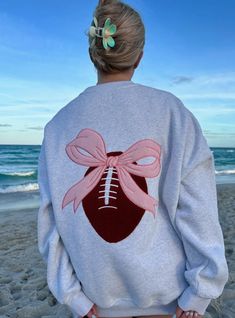 a woman standing on the beach wearing a sweatshirt with a football and pink bow tie