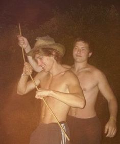 two young men standing next to each other in shorts and trunks, one holding a stick