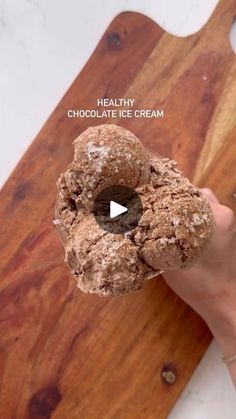 a person holding a chocolate ice cream doughnut on top of a wooden cutting board