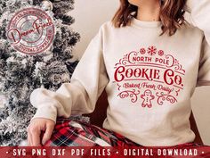 a woman sitting in front of a christmas tree wearing a sweater that says north pole cookie co