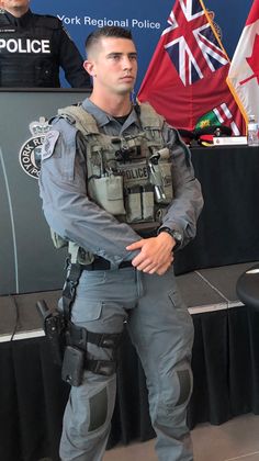 a police officer standing in front of a podium with flags behind him and another uniformed man sitting at the back